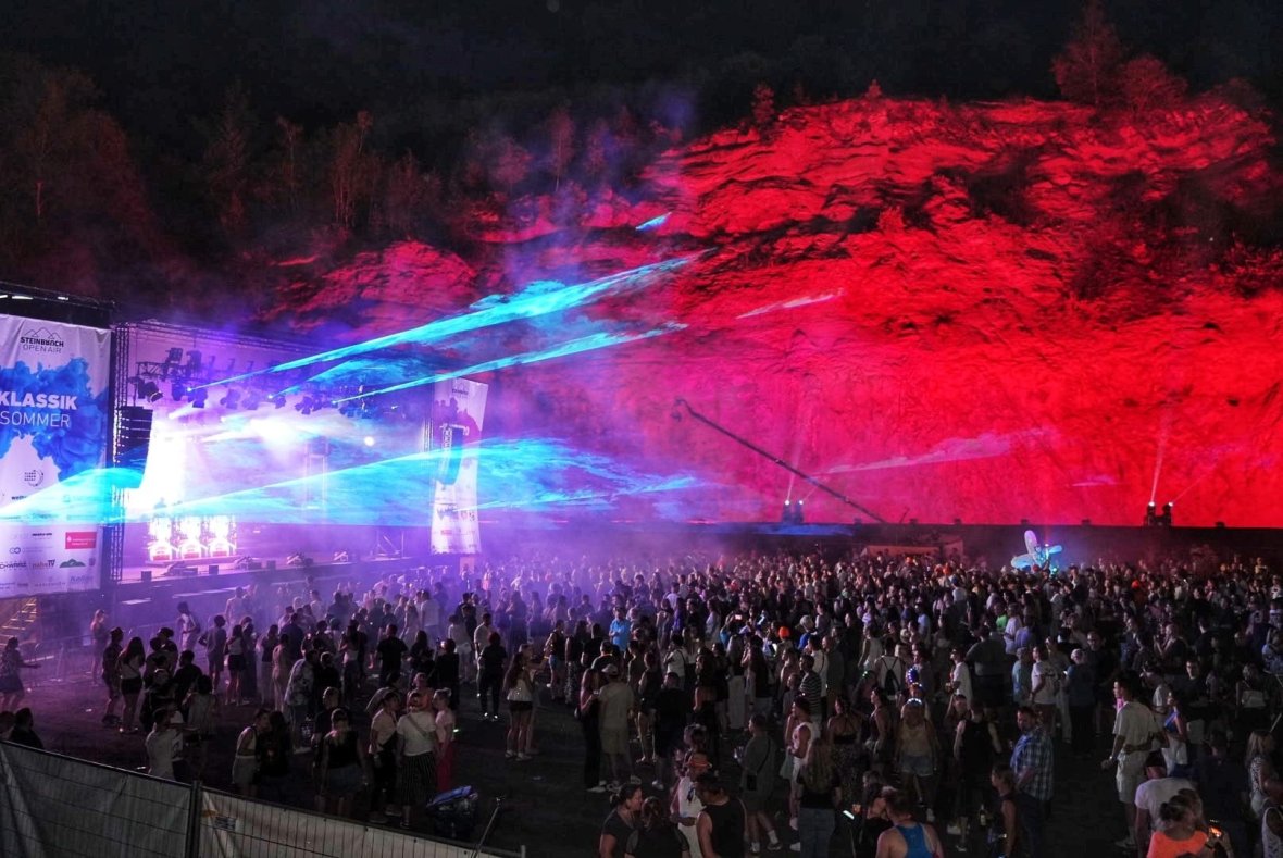 Electro-Love - rot beleuchtete Felswand Große Menschenmenge vor einer rot beleuchteten Felswand bei Nacht mit blauen Lichtstrahlen und Bühnenaufbau links