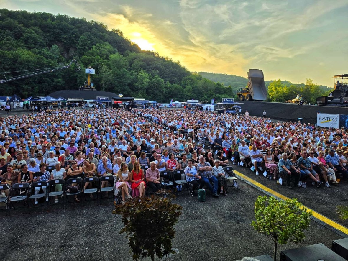 Klassik Sommer - Große Menschenmenge sitzt auf Stühlen im Freien vor einer Bühne mit Baggern im Hintergrund bei Sonnenuntergang Große Menschenmenge sitzt auf Stühlen im Freien vor einer Bühne mit Baggern im Hintergrund bei Sonnenuntergang
