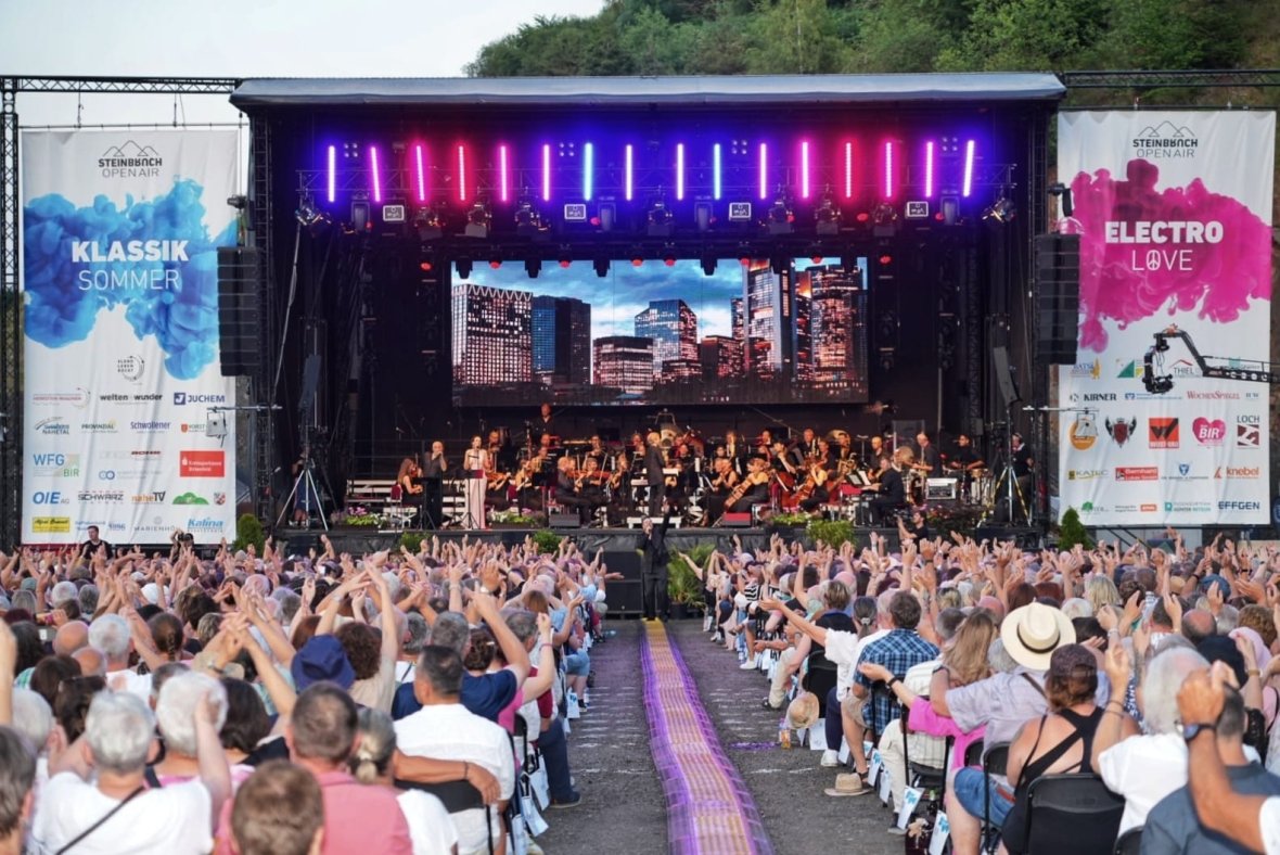 Klassik Sommer - Open-Air-Bühne mit Orchester und großem Publikum vor der Bühne bei Tageslicht Open-Air-Bühne mit Orchester und großem Publikum vor der Bühne bei Tageslicht