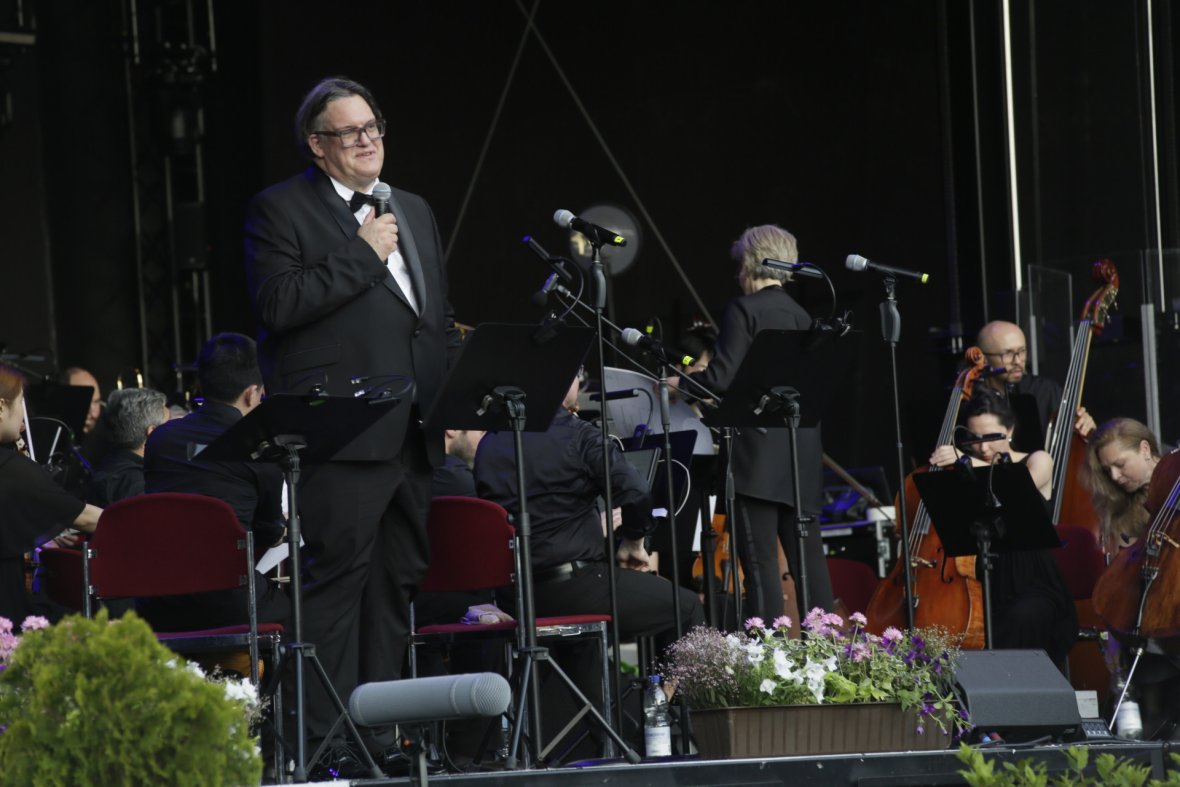 Klassik Sommer - Mann im Anzug mit Mikrofon vor einem Orchester auf einer Bühne mit Blumen im Vordergrund Mann im Anzug mit Mikrofon vor einem Orchester auf einer Bühne mit Blumen im Vordergrund