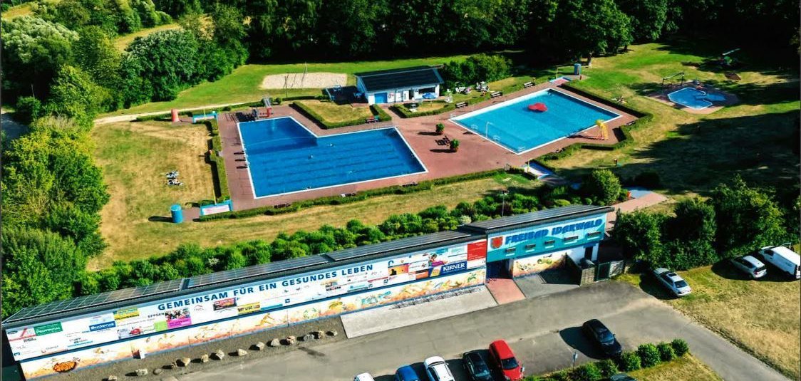 Luftaufnahme eines Freibads mit zwei Schwimmbecken, umgeben von Liegewiesen und Bäumen, davor ein Parkplatz mit mehreren Autos