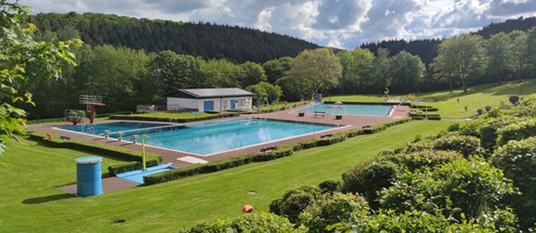 die beiden Schwimmbecken mit Sprungtürmen, Sprudler sowie Rutsche mit Betriebsgebäude (Bademeistergebäude) des Freibades Idarwald in Rhaunen mit blauer Umkleidetonne umgeben von Wiese, Bäumen und Hecken