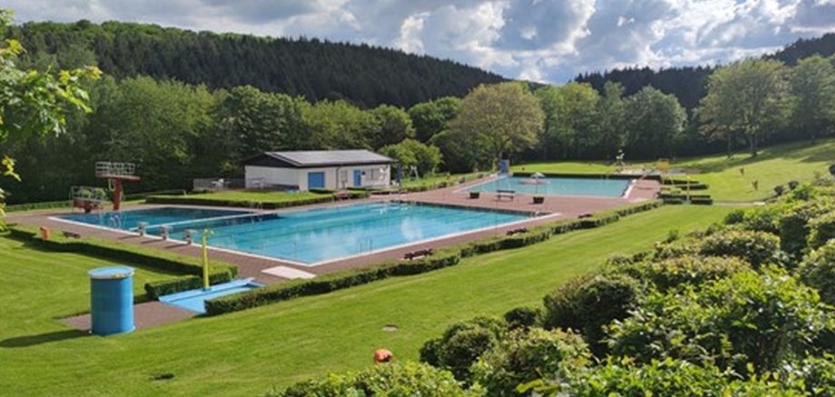 die beiden Schwimmbecken mit Sprungtürmen, Sprudler sowie Rutsche mit Betriebsgebäude (Bademeistergebäude) des Freibades Idarwald in Rhaunen mit blauer Umkleidetonne umgeben von Wiese, Bäumen und Hecken