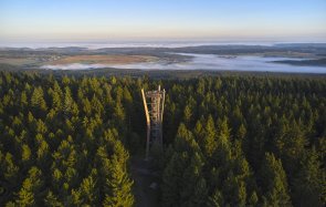 Luftbild des Idarkopfturmes inmitten von Wald. Auf dem Turm stehen ein paar Personen. In Hintergrund sind in Nebel ein paar Orte zu sehen.