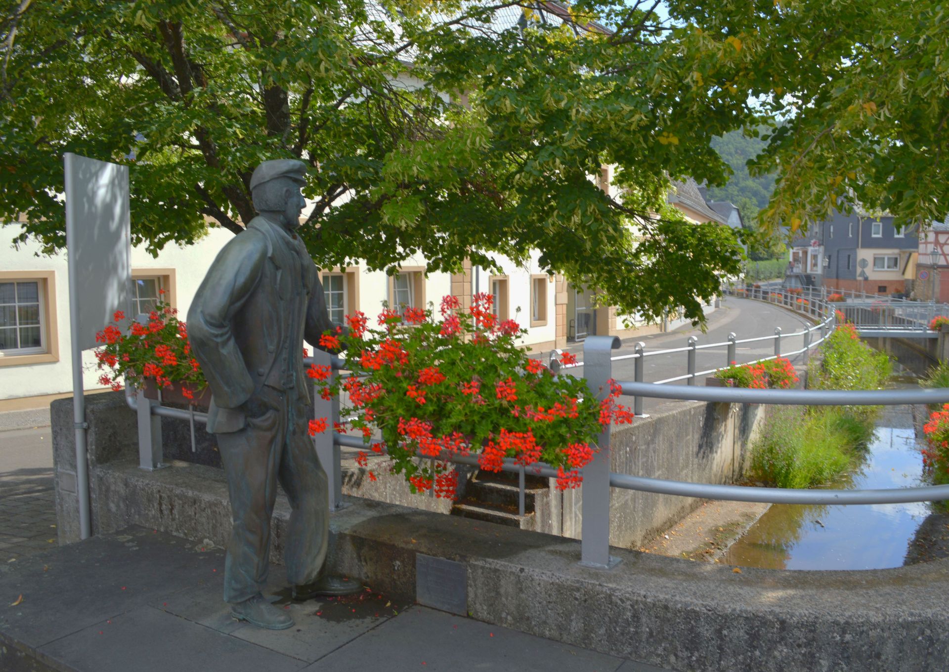 Bachspautzer in Rhaunen Statue eines Mannes mit Mütze, der in den Bach spuckt, an einem Geländer am Bach stehend an dem rote Geranien in Blumenkästen hängen
