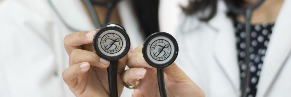 Foto Ärzte Fotografie zweier Ärztinnen (verschwommen im Hintergrund), welche beide ein Stethoskop in die Kamera halten