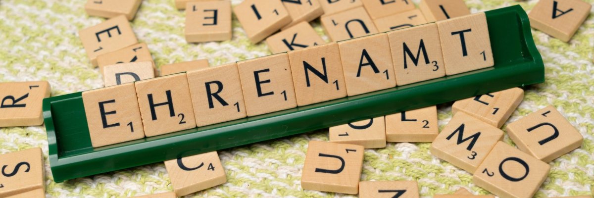 Ehrenamt scrabble Fotografie mit Scrabble-Steinen, in der Mitte das Wort Ehrenamt auf einem grünen Holzständer