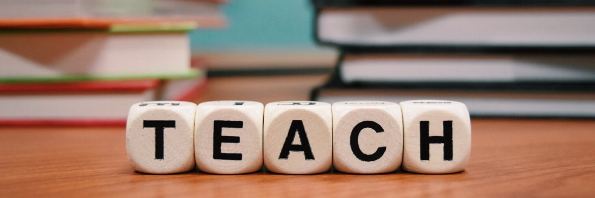 Bildung Fotografie zeigt Würfel, die das Wort Teach ergeben, im Hintergrund sieht man Bücher