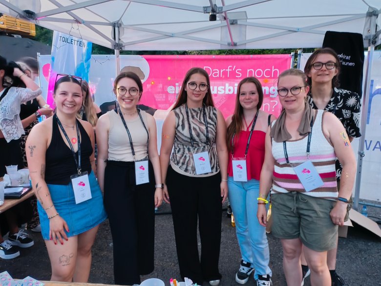 Sechs junge Frauen stehen nebeneinander vor einem Zelt mit einem pinken Banner im Hintergrund und tragen Namensschilder an schwarzen Bändern um den Hals