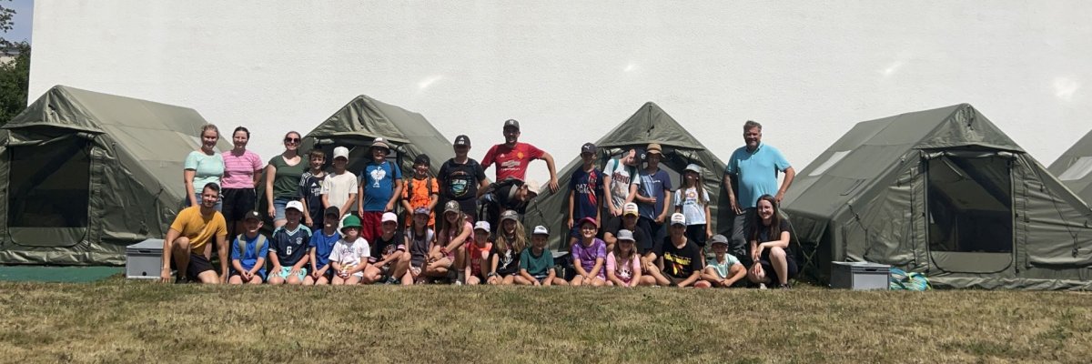 Gruppenfoto Outdoorcamp Gruppe von Erwachsenen und Kindern sitzt und steht vor mehreren Zelten auf einer Wiese vor einer großen weißen Wand