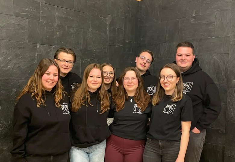 Gruppe von acht jungen Menschen in schwarzer Kleidung mit weißem Logo vor dunkler Wand