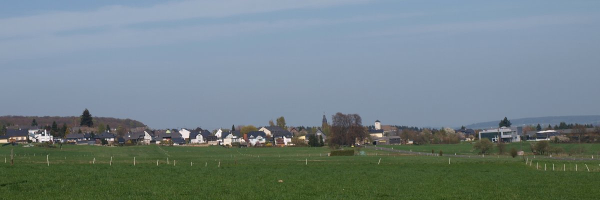 Im Vordergrund erstreckt sich eine große, grüne Wiese. Im Hintergrund sind mehrere Häuser mit weißen und dunklen Fassaden zu sehen, die am Rand der Ortsgemeinde Stipshausen stehen. Der Himmel ist hellblau mit einigen dünnen, weißen Wolken. Die Landschaft wirkt offen und weit, ohne sichtbare Bäume oder markante Erhebungen im Vordergrund.