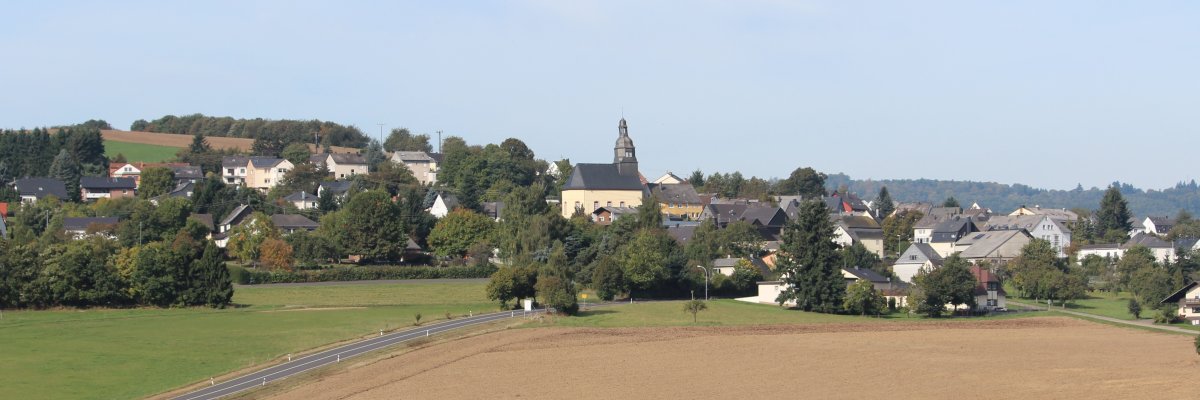 Panorama Veitsrodt Im Vordergrund erstreckt sich ein braunes Feld, dahinter eine Landstraße. Im Mittelgrund sind zahlreiche Häuser und Gebäude zu sehen, die von Bäumen und Grünflächen umgeben sind. Im Zentrum ragt eine große Kirche mit einem markanten Turm hervor. Der Hintergrund zeigt sanfte Hügel und bewaldete Flächen unter einem hellblauen Himmel.