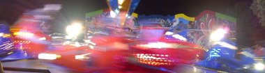 Prämienmarkt Veitsrodt Bunte Lichter und verschwommene Bewegungen dominieren das Bild. Im Vordergrund sind leuchtend rote und blaue Streifen zu sehen, die auf die schnelle Bewegung eines Fahrgeschäfts hindeuten. Im Hintergrund befindet sich eine farbenfrohe Wand mit großflächigen, abstrakten Mustern und geschwungenen Linien. Die Szene spielt sich vermutlich nachts oder in der Dämmerung ab, da der Himmel dunkel ist und die Lichter besonders hervorstechen.