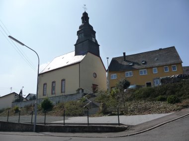 Kirche Veitsrodt Kirchgebäude mit Turm und Uhr an einer leicht ansteigenden Straße, daneben Wohnhäuser und ein Laternenmast