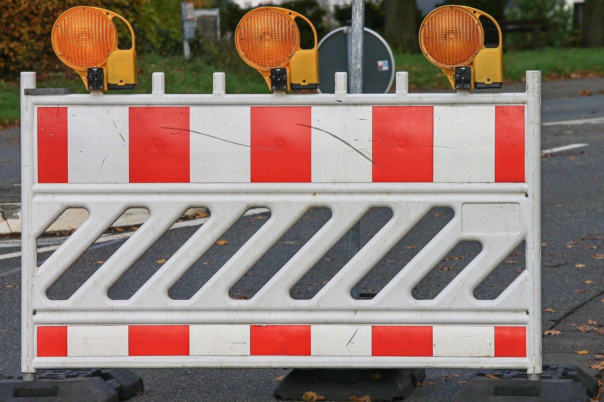 zu sehen hier ist eine Straßenabsperrung mit einem weißen gitter welches mit orangenen Streifen versehen ist und 3 orangefarbene Leuchten obendrauf hat