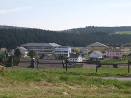 Im Hintergrund das Schulgebäude in Rhaunen mit angrenzender Idarwaldhalle sowie des Sportplatzes. Im Vordergrund einige Häuser und eine Rasenfläche: