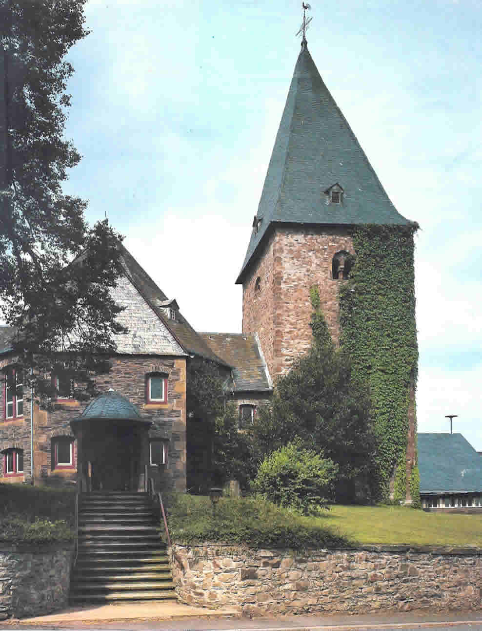 Foto Kirche Hottenbach Kirche mit steilem Schieferdach und teilweise mit Efeu bewachsenem Turm neben einem Treppenaufgang und einer Steinmauer