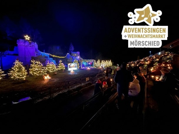 Weihnachtlich beleuchteter Außenbereich mit Tannenbäumen, einer Bühne und Publikum bei Nacht