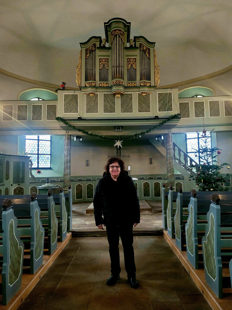 Foto Thomas Diedrich Person mit dunkler Kleidung steht in der Mitte eines Kircheninnenraums vor einer Orgel und neben einem geschmückten Weihnachtsbaum