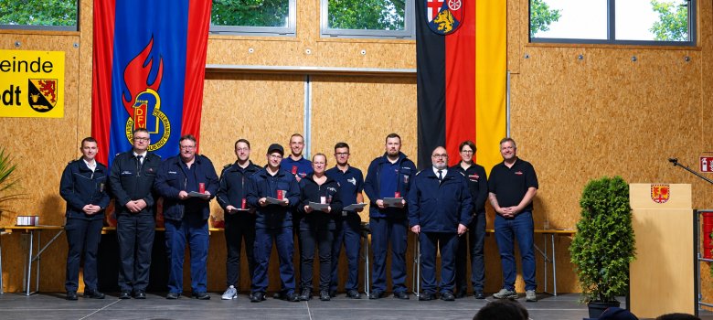 Gruppe von Männern in Uniform auf einer Bühne vor zwei großen Fahnen und einem Rednerpult mit Wappen