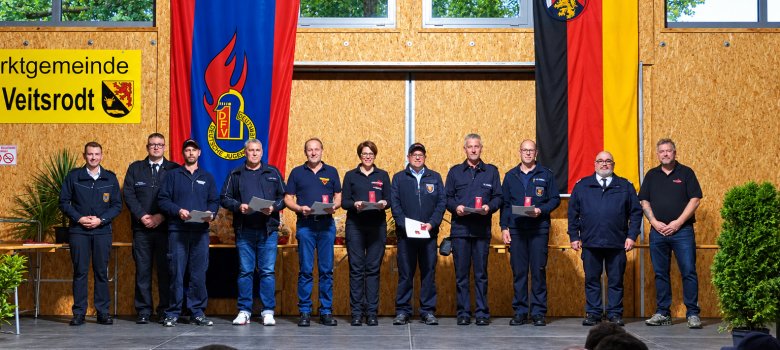 Zehn Personen in dunkler Kleidung stehen auf einer Bühne vor zwei großen Fahnen und einem gelben Schild mit der Aufschrift 'Marktgemeinde Veitsrodt'
