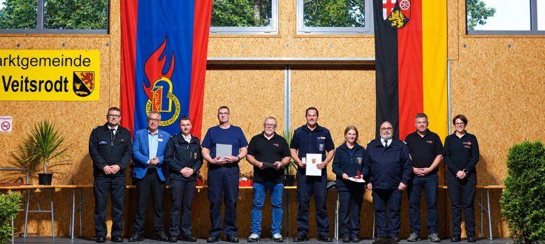 35 aktive Jahre in der Feuerwehr Zehn Personen stehen auf einer Bühne vor zwei großen Fahnen und einem Schild mit der Aufschrift 'Marktgemeinde Veitsrodt'