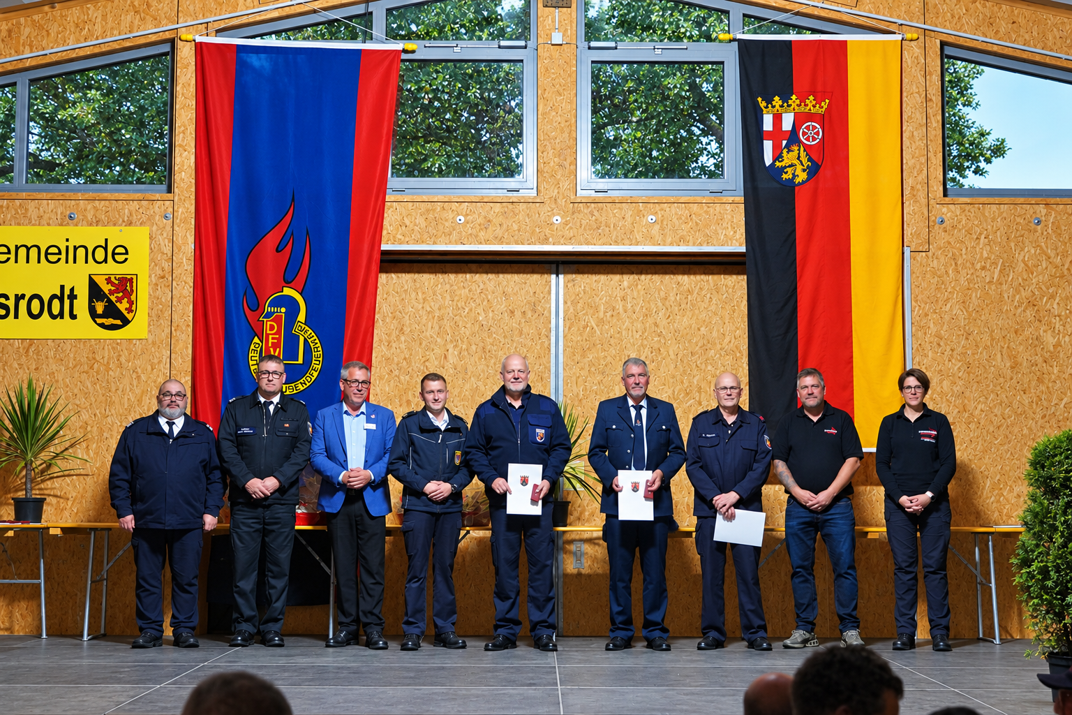 Acht Personen in Uniform und Anzug stehen auf einer Bühne vor zwei großen Fahnen und einer gelben Gemeindetafel