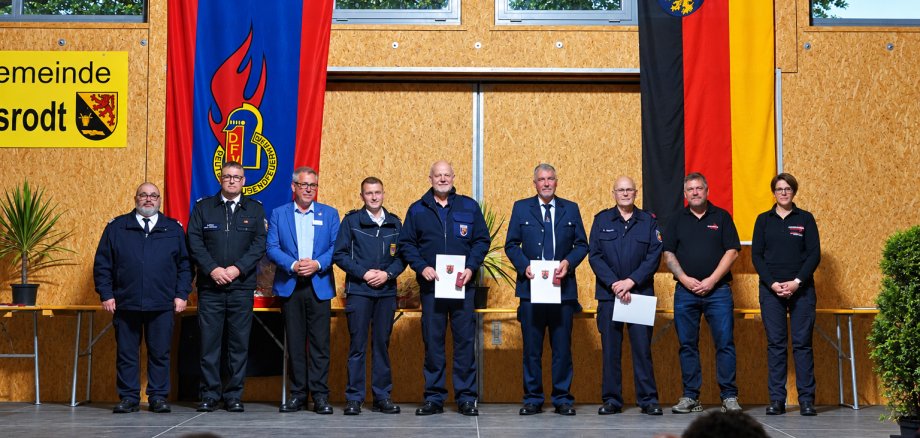 45 aktive Jahre in der Feuerwehr Acht Personen in Uniform und Anzug stehen auf einer Bühne vor zwei großen Fahnen und einer gelben Gemeindetafel