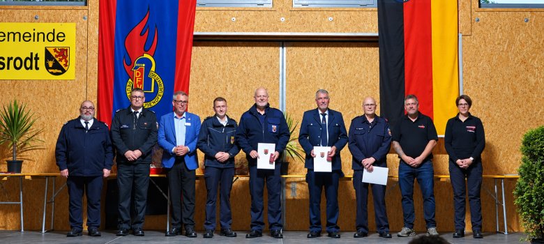45 aktive Jahre in der Feuerwehr Acht Personen in Uniform und Anzug stehen auf einer Bühne vor zwei großen Fahnen und einer gelben Gemeindetafel