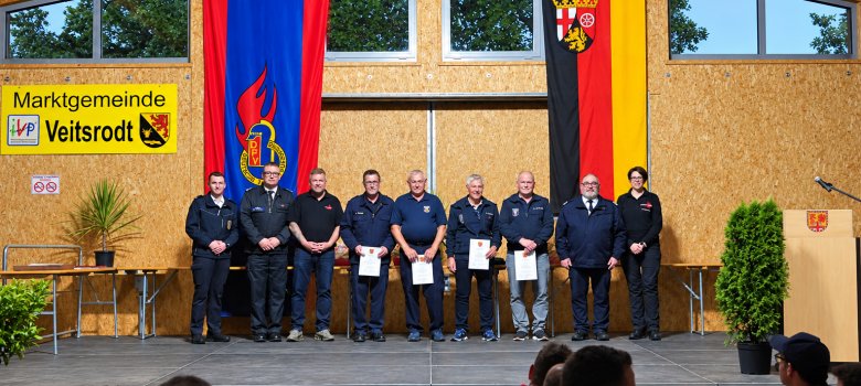 Entlassungen aus dem Feuerwehrdienst Acht Personen in Uniform stehen auf einer Bühne vor zwei großen Fahnen und einem Schild mit der Aufschrift 'Marktgemeinde Veitsrodt'