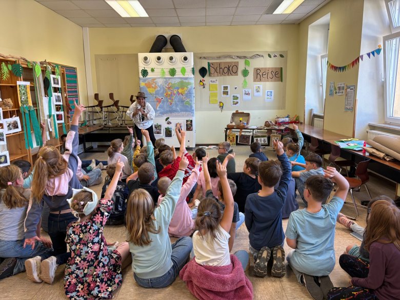 Kinder sitzen auf dem Boden eines Klassenraums und heben die Hände, während eine Person vorne mit Hut spricht. In einem Klassenzimmer sitzen zahlreiche Kinder auf dem Boden und heben die Hände. Im Vordergrund sind die Kinder von hinten zu sehen, sie tragen bunte Alltagskleidung. Im Hintergrund steht eine erwachsene Person mit Hut und Mikrofon vor einer Weltkarte und mehreren Plakaten, auf denen unter anderem 'Schoko Reise' steht. Die Wände sind mit Fotos, Girlanden und weiteren Dekorationen geschmückt. Rechts stehen Tische und Stühle, auf einem Tisch liegen zusammengerollte Matten oder Poster.