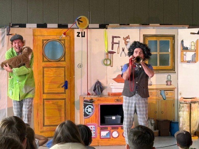 Zwei Schauspieler in bunter Kleidung auf einer Bühne mit einer Holzhaustür und einem gemalten Fenster, einer hält ein Stofftier, der andere spielt Trompete, Publikum im Vordergrund Im Vordergrund stehen zwei Personen auf einer Bühne. Die linke Person trägt ein hellgrünes Hemd, eine karierte Hose und eine Mütze und hält einen braunen Hund im Arm. Die rechte Person trägt eine Weste, eine karierte Hose und eine schwarze Lockenperücke und spielt eine Trompete. Im Hintergrund ist eine bemalte Kulisse mit einer Tür, einem Fenster, Küchenelementen und verschiedenen Gegenständen zu sehen. Vor der Bühne sitzen mehrere Kinder und schauen zu.