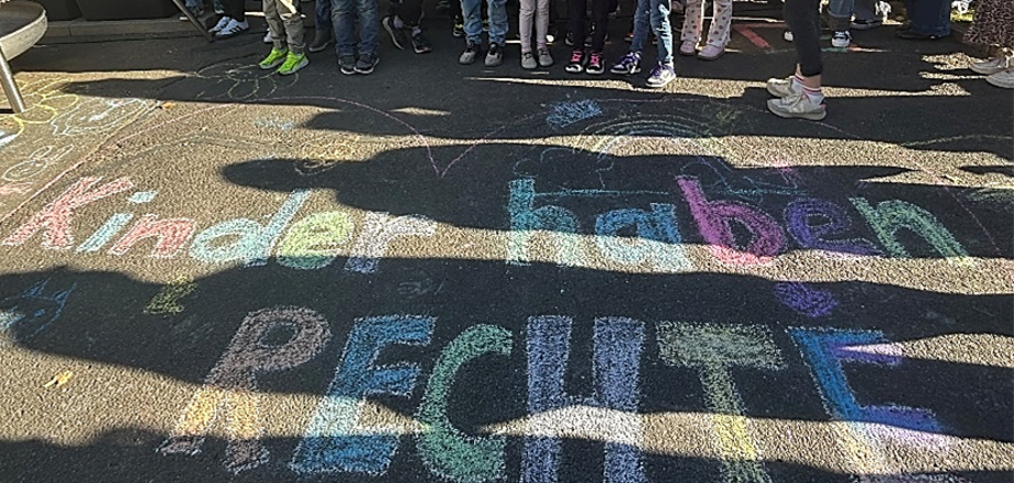 Kinder stehen auf einem Asphaltboden, auf dem mit bunter Kreide die Worte 'kinder haben RECHTE' geschrieben sind Bunte Kreidebuchstaben auf dunklem Asphalt bilden den Schriftzug 'Kinder haben RECHTE'. Im oberen Bildbereich stehen mehrere Kinder dicht nebeneinander, ihre Beine und Schuhe sind sichtbar. Sonnenlicht und Schatten von Personen fallen quer über die Kreidezeichnung.