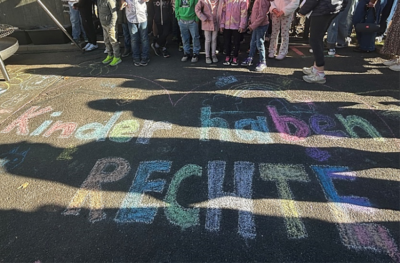 Kinder stehen auf einem Asphaltboden, auf dem mit bunter Kreide die Worte 'kinder haben RECHTE' geschrieben sind Bunte Kreidebuchstaben auf dunklem Asphalt bilden den Schriftzug 'Kinder haben RECHTE'. Im oberen Bildbereich stehen mehrere Kinder dicht nebeneinander, ihre Beine und Schuhe sind sichtbar. Sonnenlicht und Schatten von Personen fallen quer über die Kreidezeichnung.