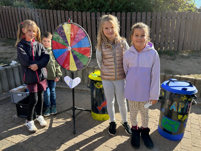 Vier Kinder stehen neben einem bunten Glücksrad und zwei bemalten Mülltonnen auf einem gepflasterten Hof vor einem Holzzaun Vier Kinder stehen im Freien auf einem gepflasterten Weg. Zwei von ihnen stehen direkt neben einem bunten Glücksrad, das auf einem Ständer befestigt ist. Die beiden anderen Kinder stehen etwas abseits, nahe einer niedrigen Mauer. Rechts neben dem Glücksrad befinden sich zwei große Mülltonnen mit farbigen Aufklebern und der Aufschrift 'PAPIER'. Im Hintergrund ist ein Holzzaun und grüne Vegetation zu sehen. Die Kinder tragen Jacken und lächeln.
