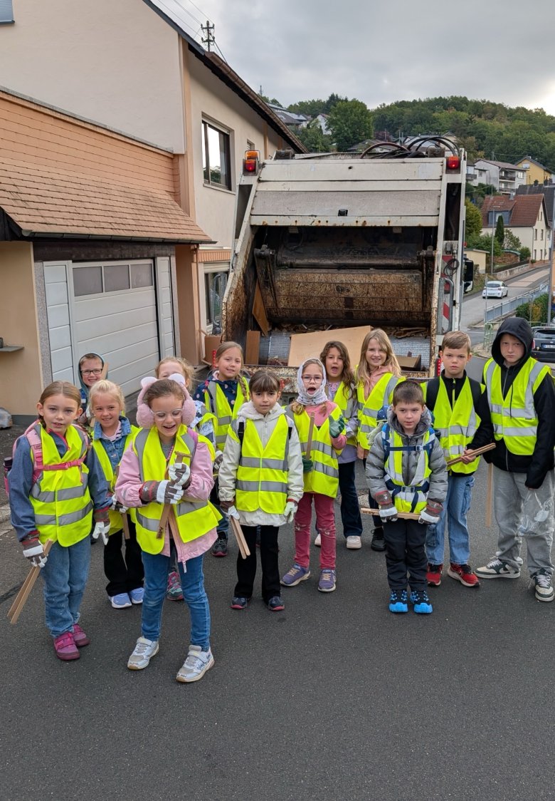 Gruppe von Kindern mit gelben Warnwesten steht vor einem Müllwagen auf einer Straße in einem Wohngebiet Eine Gruppe von Kindern steht auf einer Straße vor einem Müllfahrzeug. Alle tragen leuchtend gelbe Warnwesten und halten Greifzangen in den Händen. Im Hintergrund sind Wohnhäuser und eine hügelige Landschaft mit Bäumen zu sehen. Die Kinder blicken in Richtung Kamera.